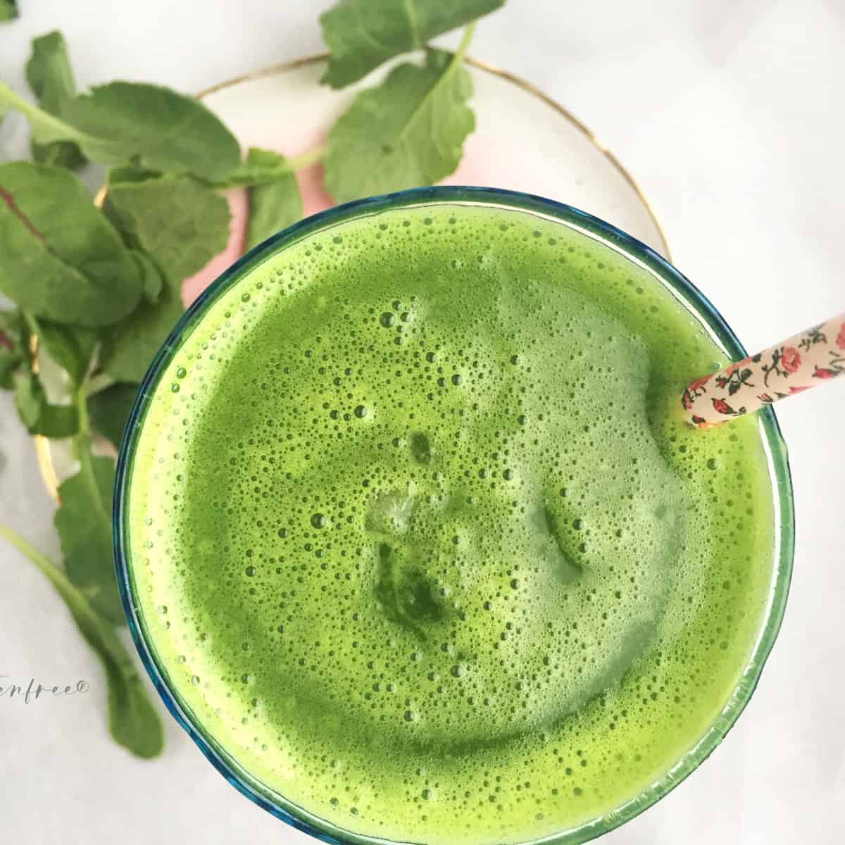 short glass of green juice with floral straw on top of a marble table with a pink plate and mixed green garnish